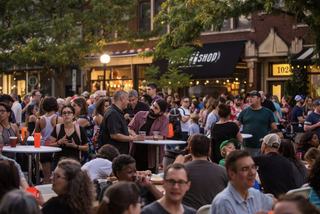 Crowds gather at Oaktoberfest in Oakland, enjoying food, drinks, and live music at outdoor tables along a tree-lined street with local storefronts in the background.