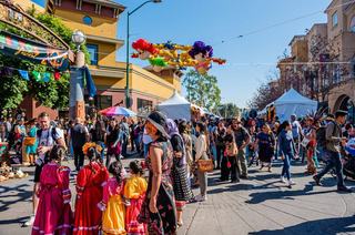 A vibrant street scene at Oakland’s Día de los Muertos Festival in Fruitvale, with families, dancers in colorful traditional dresses, artisan tents, and decorative papel picado banners overhead.