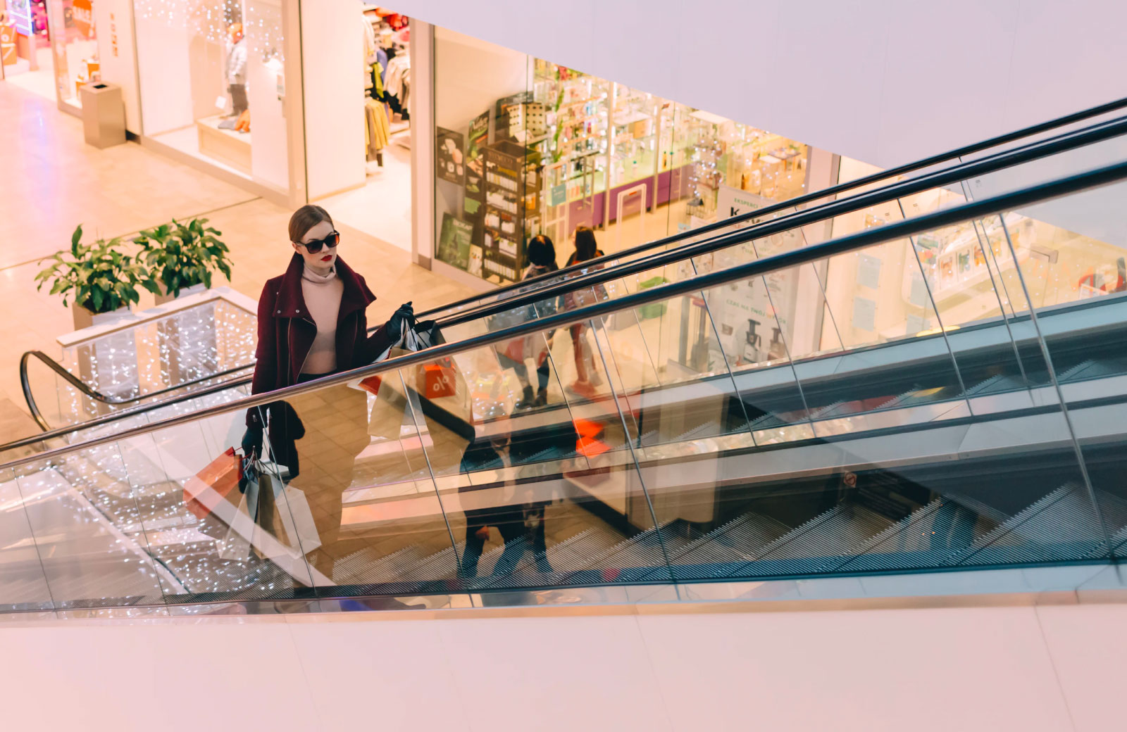 woman in jacket and shades holding shopping bags on an mall escalator
