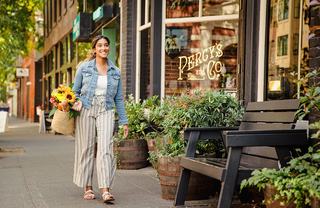 A woman walks along a leafy Ballard sidewalk past Percy’s & Co., carrying flowers and a market bag, with neighborhood shops, planters, and benches creating a relaxed, walkable street scene.