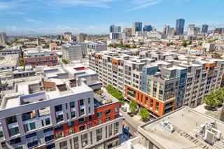 An aerial, high-angle view of Fourth Street East apartments in Oakland's Jack London Square. The modern, mid-rise residential buildings feature a mix of grey, red, and burnt-orange facades with industrial-chic architecture and private balconies. In the background, the downtown Oakland skyline rises under a clear blue sky, illustrating the neighborhood's close proximity to the city center and its mix of residential and urban landscapes.