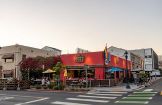 A bright red restaurant in Hillcrest with patio seating, colorful umbrellas, and a pride flag, surrounded by flowering vines and people dining along a lively neighborhood corner at dusk.