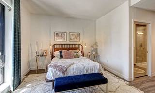A bohemian-decorated bedroom in one of our one-bedroom apartments in downtown Stamford. There is a chevron rug placed over plank flooring, nightstands with candles, and a neutral color palette. You can see a tall window bringing in natural light and the entrance to the spacious bathroom.