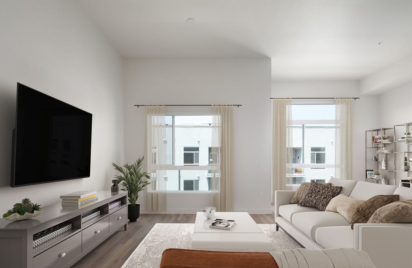Modern townhome living room with large windows framed by sheer beige curtains, a white sofa with patterned throw pillows, a low white coffee table, wall-mounted flat-screen TV, and neutral-toned decor on hardwood floors.