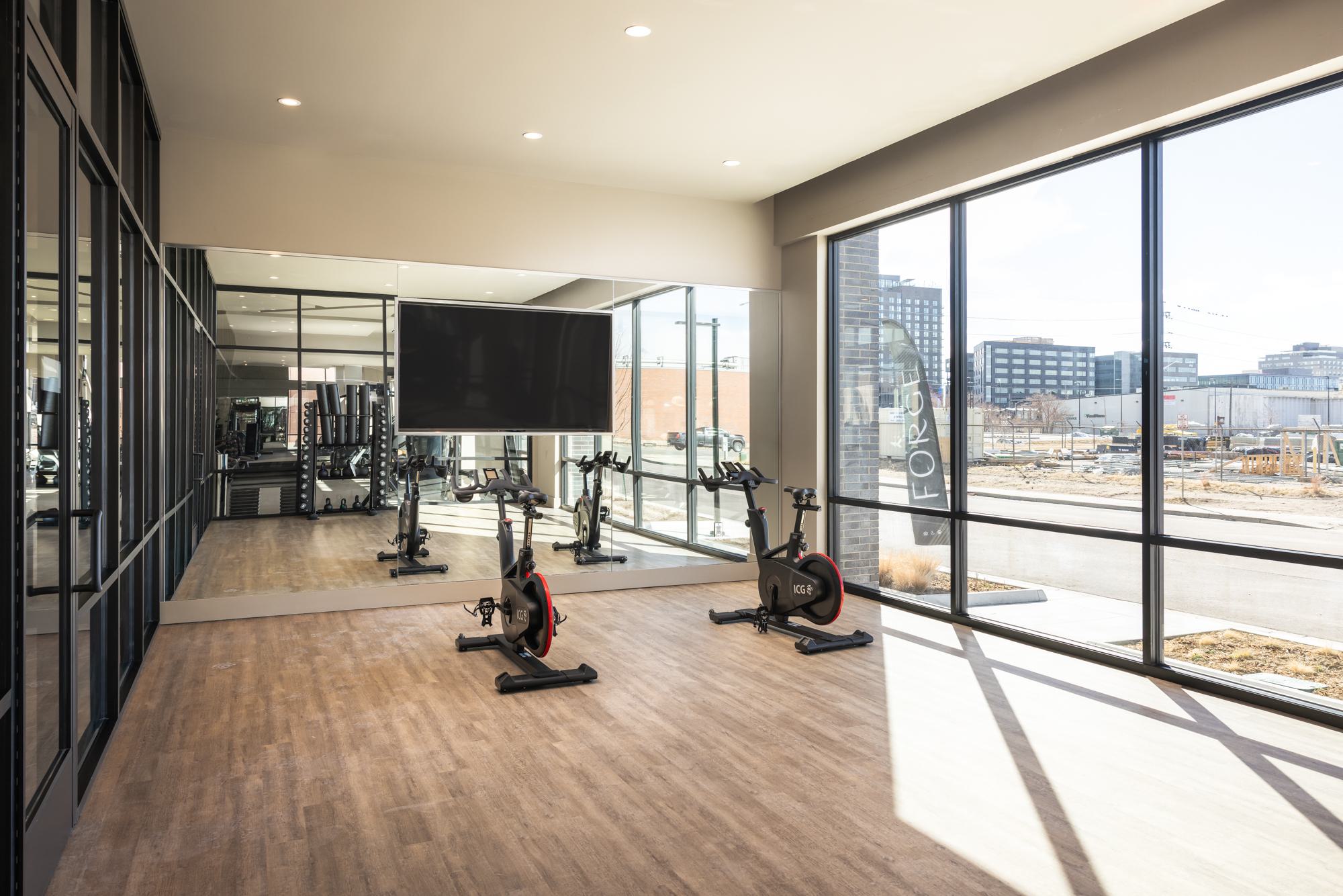 Bright and modern fitness-on-demand studio at Forge Apartments in RiNo, Denver, featuring two stationary bikes, mirrored wall, wood-look flooring, and a large screen for virtual workouts, with floor-to-ceiling windows overlooking the city.