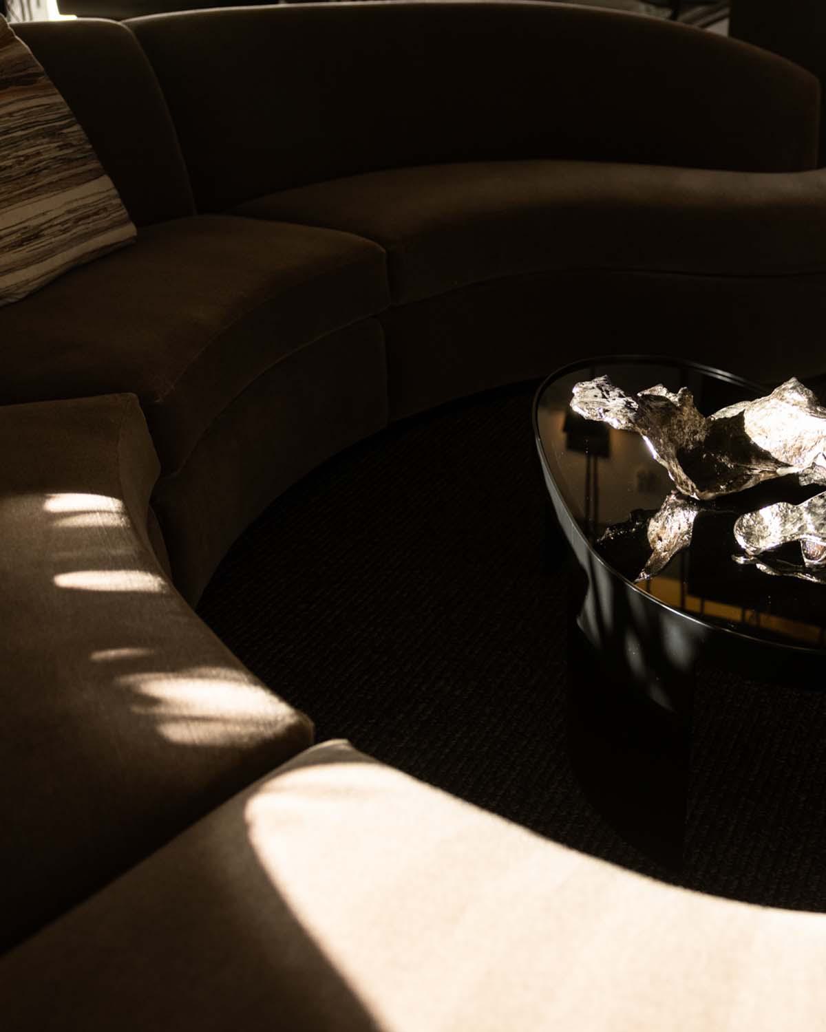 Minimalist tabletop scene in an Alloy apartment, featuring a curved cream-colored chair, a white round table, and a design book opened to pages with rustic interior imagery. A modern succulent arrangement with driftwood casts soft shadows in the sunlit corner, creating a calm, artful vignette.