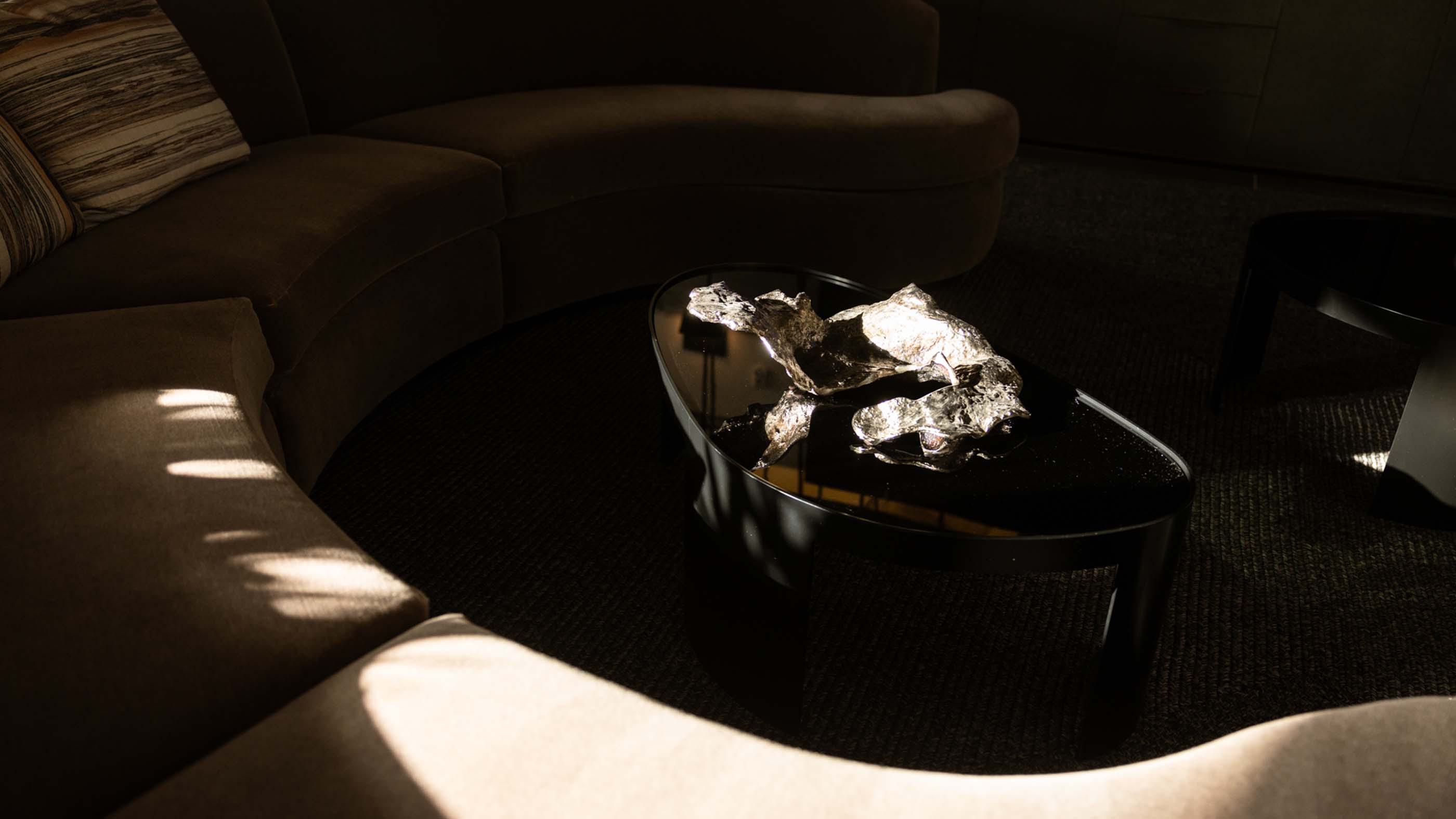 Minimalist tabletop scene in an Alloy apartment, featuring a curved cream-colored chair, a white round table, and a design book opened to pages with rustic interior imagery. A modern succulent arrangement with driftwood casts soft shadows in the sunlit corner, creating a calm, artful vignette.