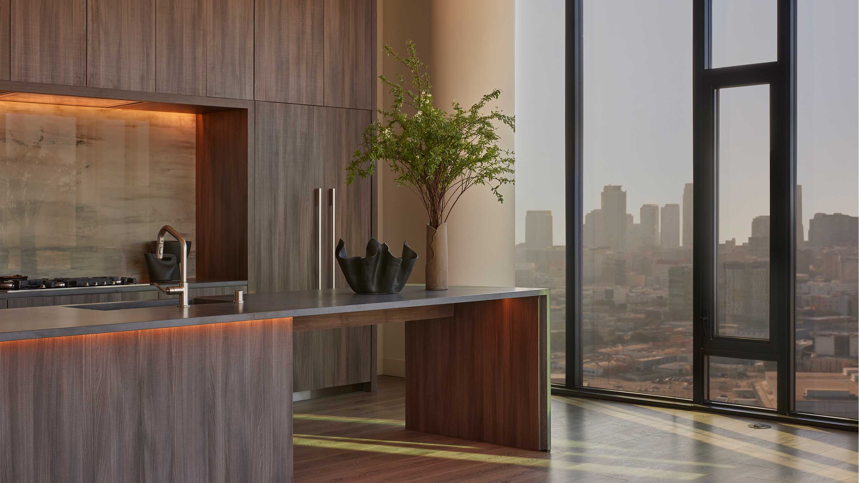 Penthouse living space at Alloy LA, featuring sculptural furnishings, a textured accent wall, and floor-to-ceiling windows framing panoramic sunset views of the downtown Los Angeles skyline.