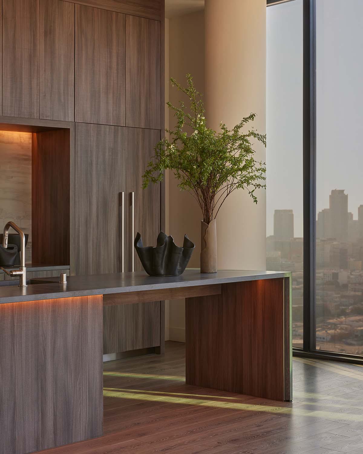 Penthouse living space at Alloy LA, featuring sculptural furnishings, a textured accent wall, and floor-to-ceiling windows framing panoramic sunset views of the downtown Los Angeles skyline.