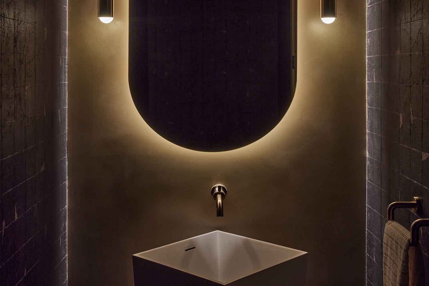 Moody, modern powder room in an Alloy penthouse, featuring a sculptural sink, wall-mounted faucet, dark tile walls, and a softly backlit oval mirror.
