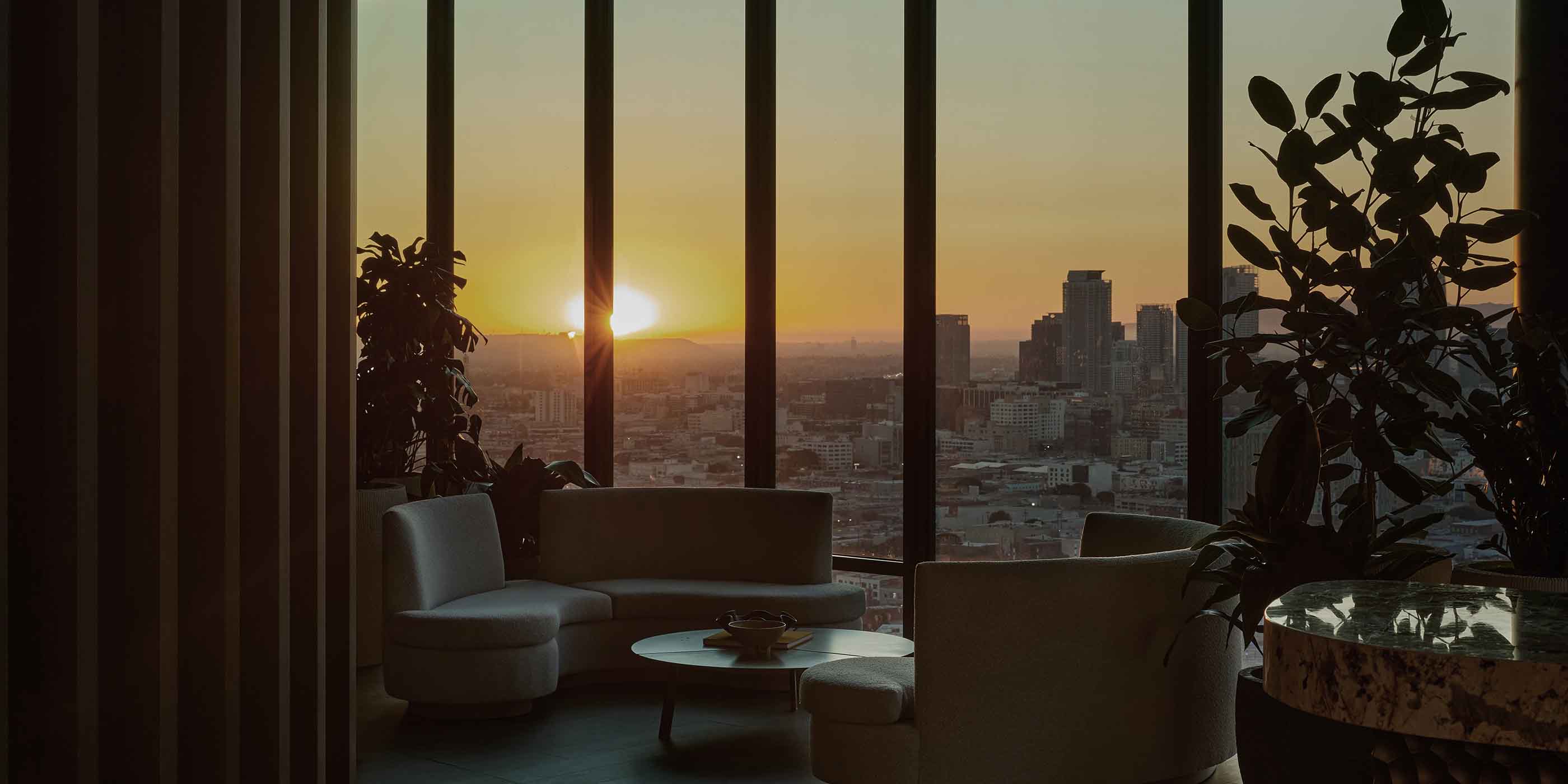 Sunset view over downtown Los Angeles seen through floor-to-ceiling windows in a modern high-rise lounge, featuring curved seating, leafy indoor plants, and a stone-top table in the foreground.