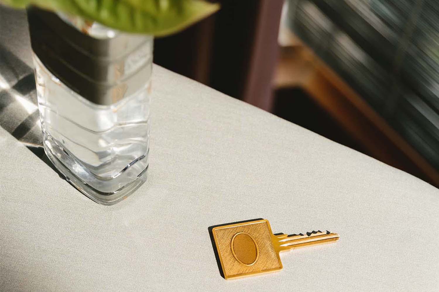 Golden key on a light fabric surface beside a glass vase in sunlight.