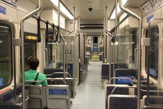 Interior of a Seattle Link light rail train, showing rows of blue seats, silver handrails and overhead grab straps, wide center aisle, accessibility signage, and a rider seated near the front of the car.