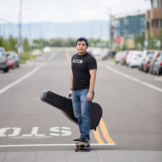 male Student in jeans and black t-shirt holding Guitar Case walking down the street - Sol at West Village Apts