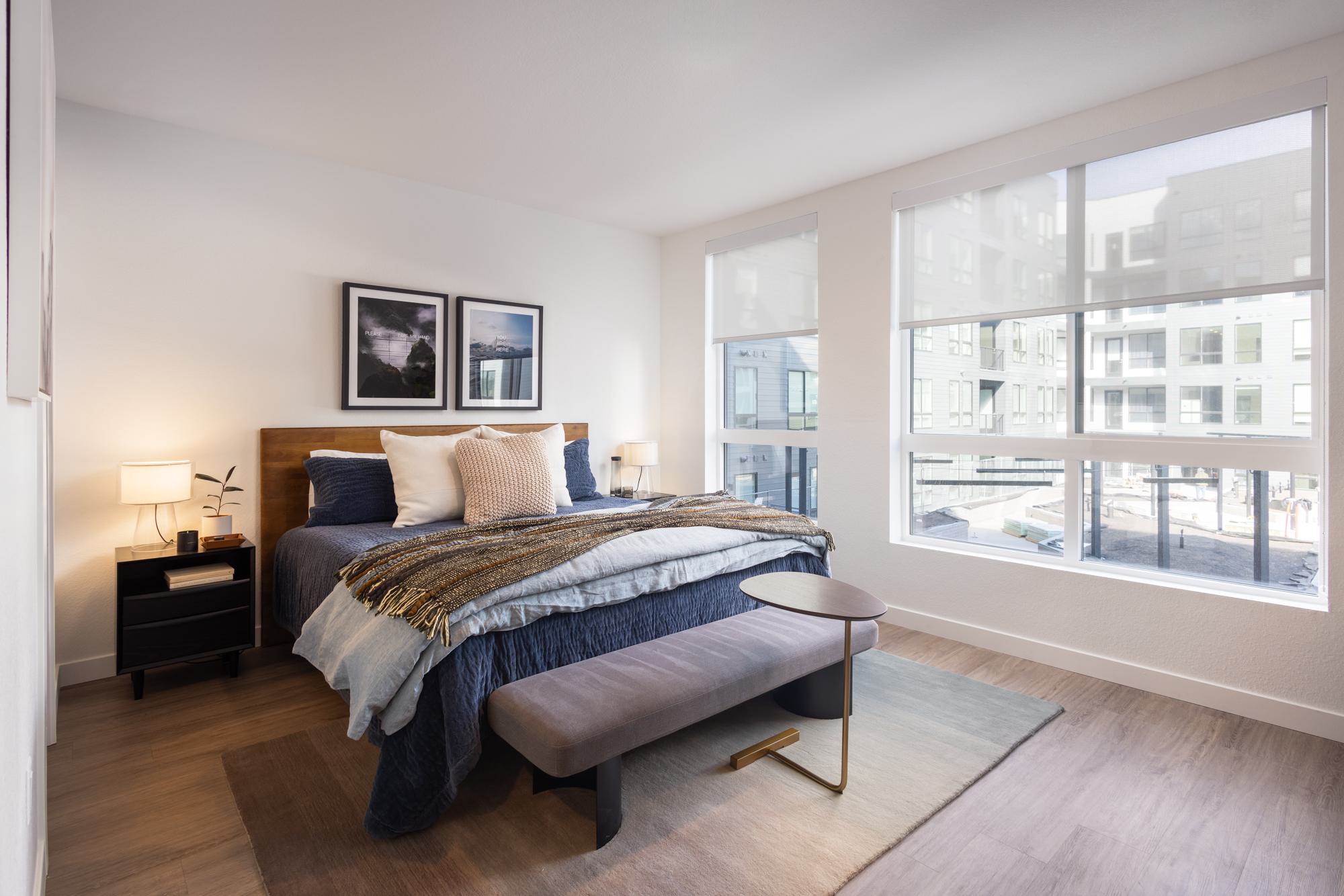 Studio bedroom with oversized corner windows, a wood-frame bed with layered bedding, and a soft bench at the foot of the bed on a neutral area rug
