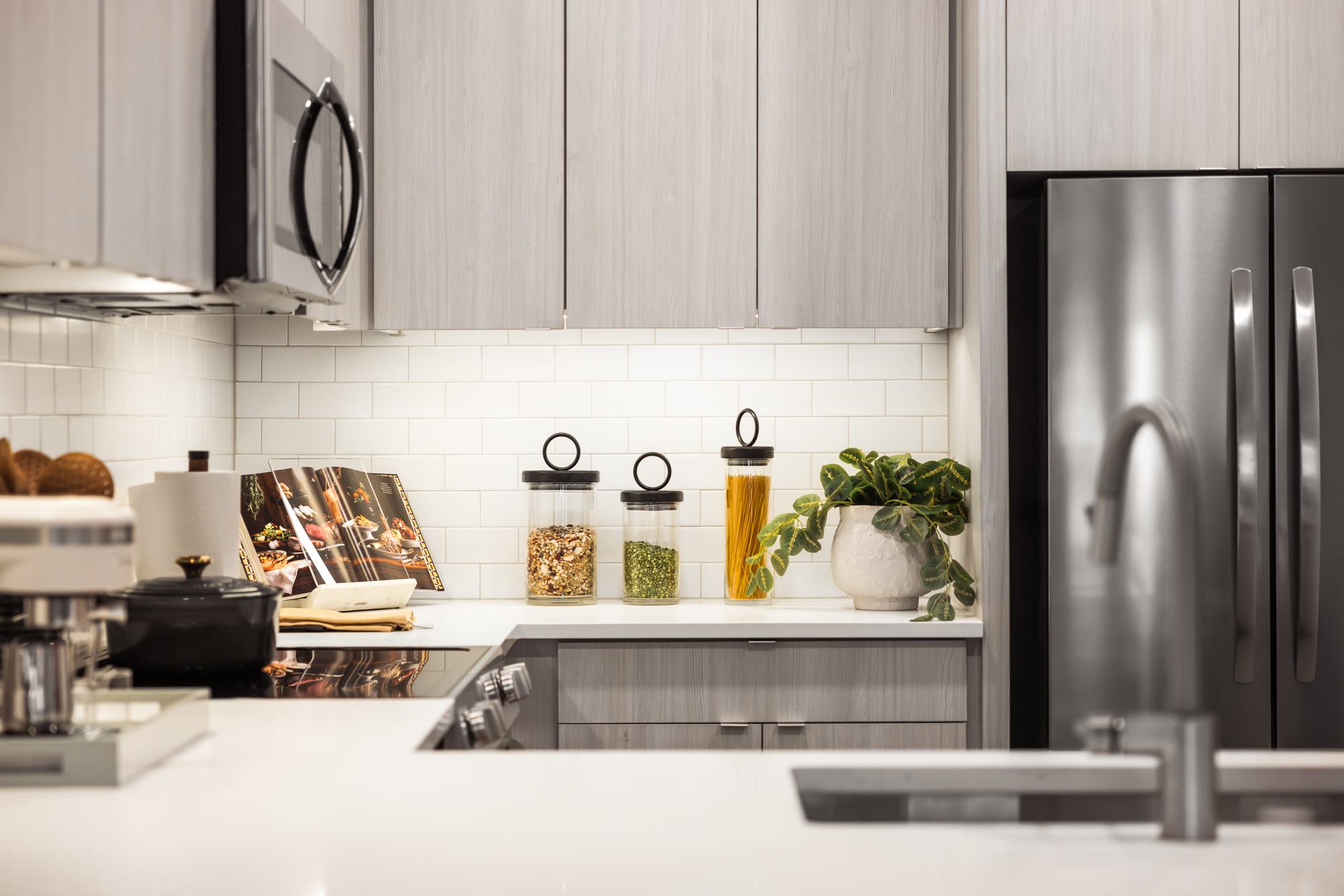 Modern kitchen at Forge Apartments in RiNo, Denver, featuring light gray cabinetry, white quartz countertops, a white subway tile backsplash, and decorative touches including a plant, glass canisters, and an open cookbook.