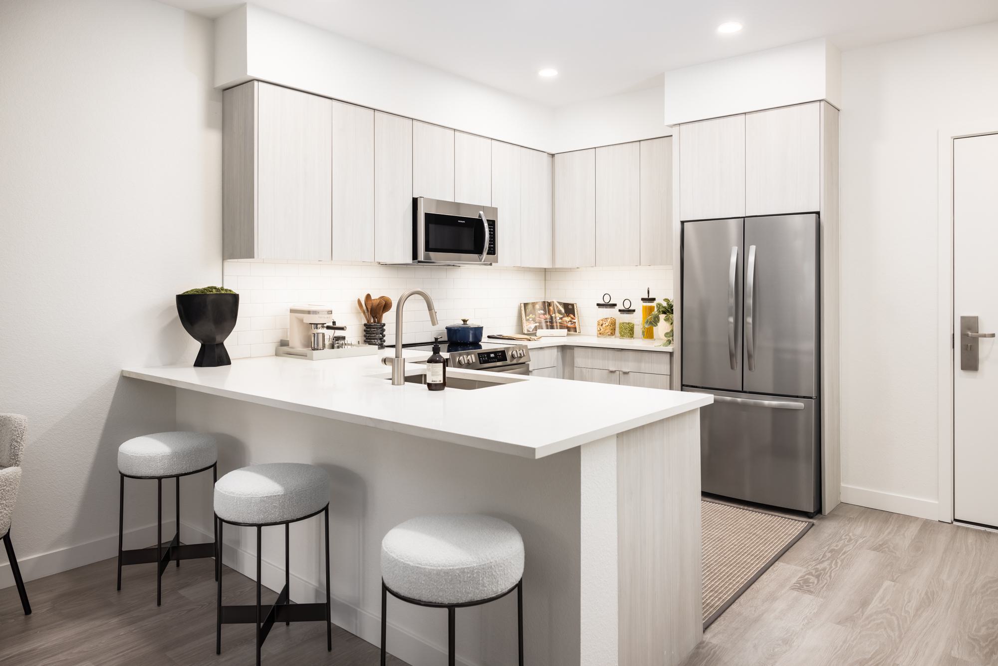 Kitchen featuring a cool-toned color scheme with light gray cabinetry, white quartz countertops, stainless steel appliances, and minimalist decor, including modern barstools and a sculptural black planter.