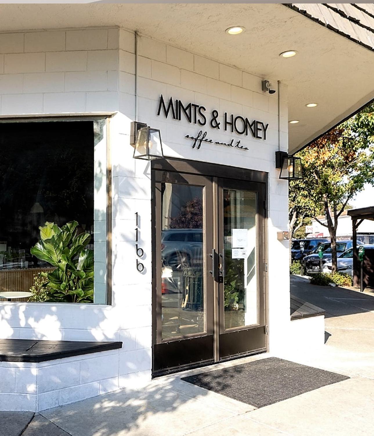 Street view of Mints & Honey, a minimalist coffee and tea shop with a crisp white tile exterior, black-framed glass doors, and leafy plants in downtown Burlingame, CA.