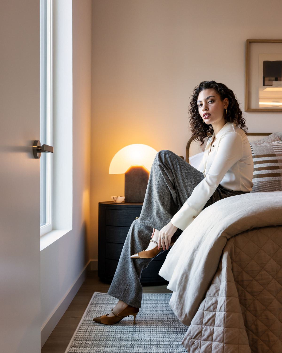 A woman sits on the edge of a bed in a light-filled room with modern styling.