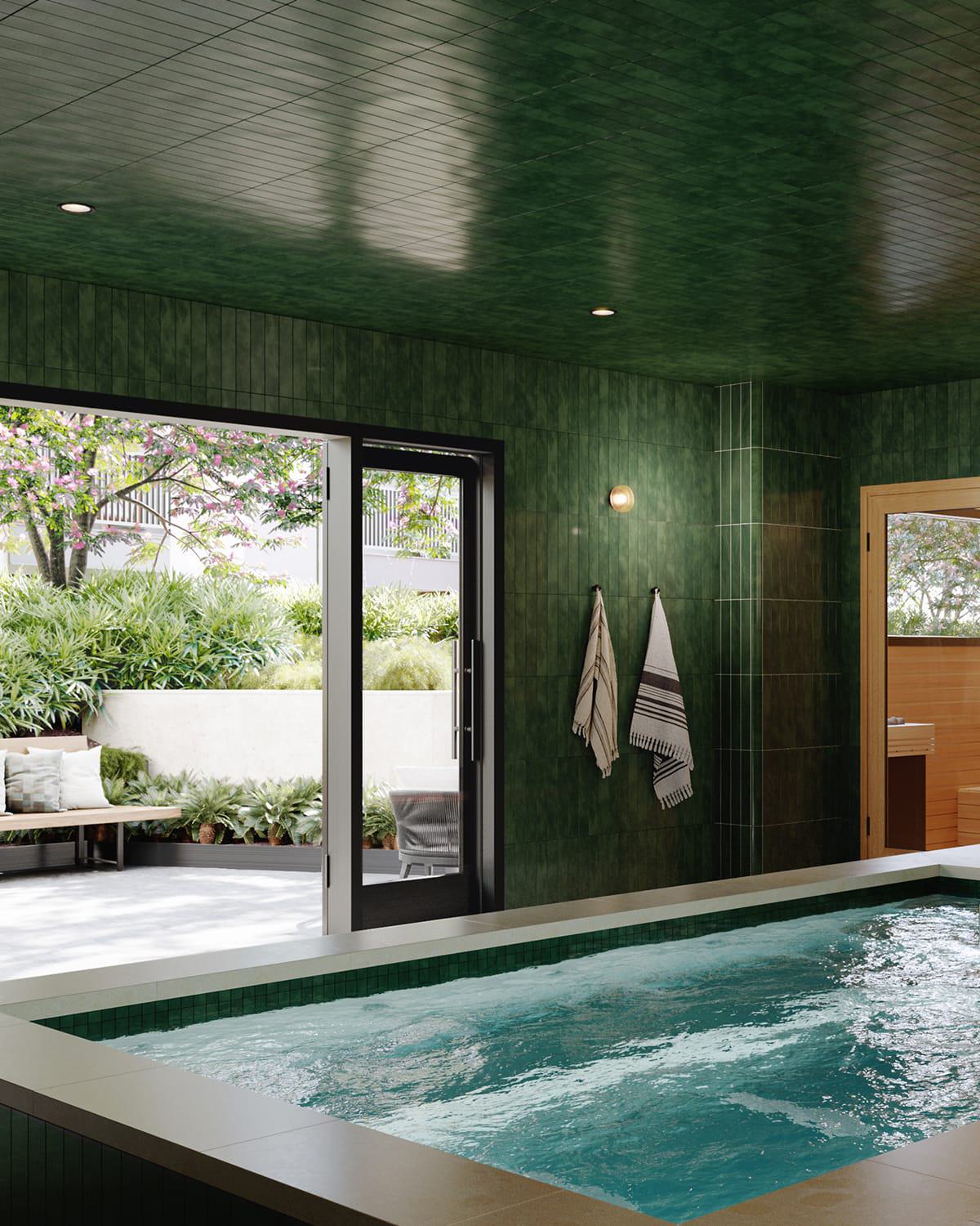 A serene indoor spa features a rectangular soaking pool framed by rich green tile walls and wood-accented sauna rooms. Floor-to-ceiling glass doors open to a tranquil outdoor lounge with blooming trees, cushioned benches, and lush landscaping.