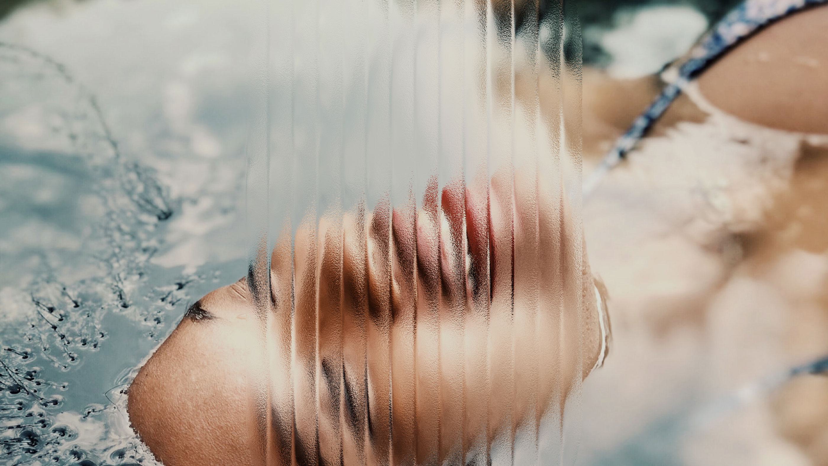 A relaxed person floats on top of the water, partially obscured by ribbed translucent glass, creating a soft, abstract, and dreamlike effect.