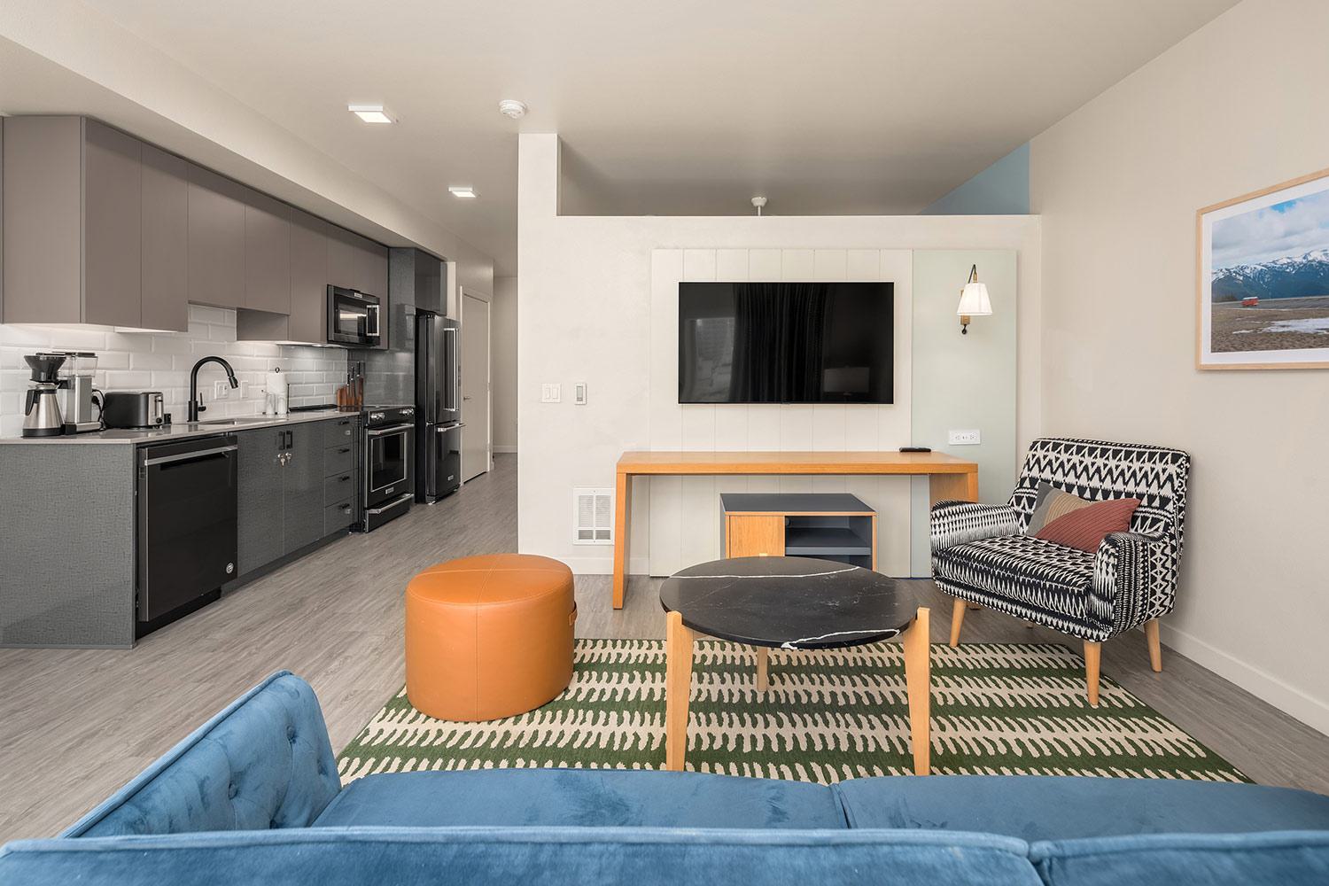  Contemporary furnished apartment living room with a patterned armchair, round ottoman, flat-screen TV, and an open kitchen featuring sleek cabinetry and modern appliances.
