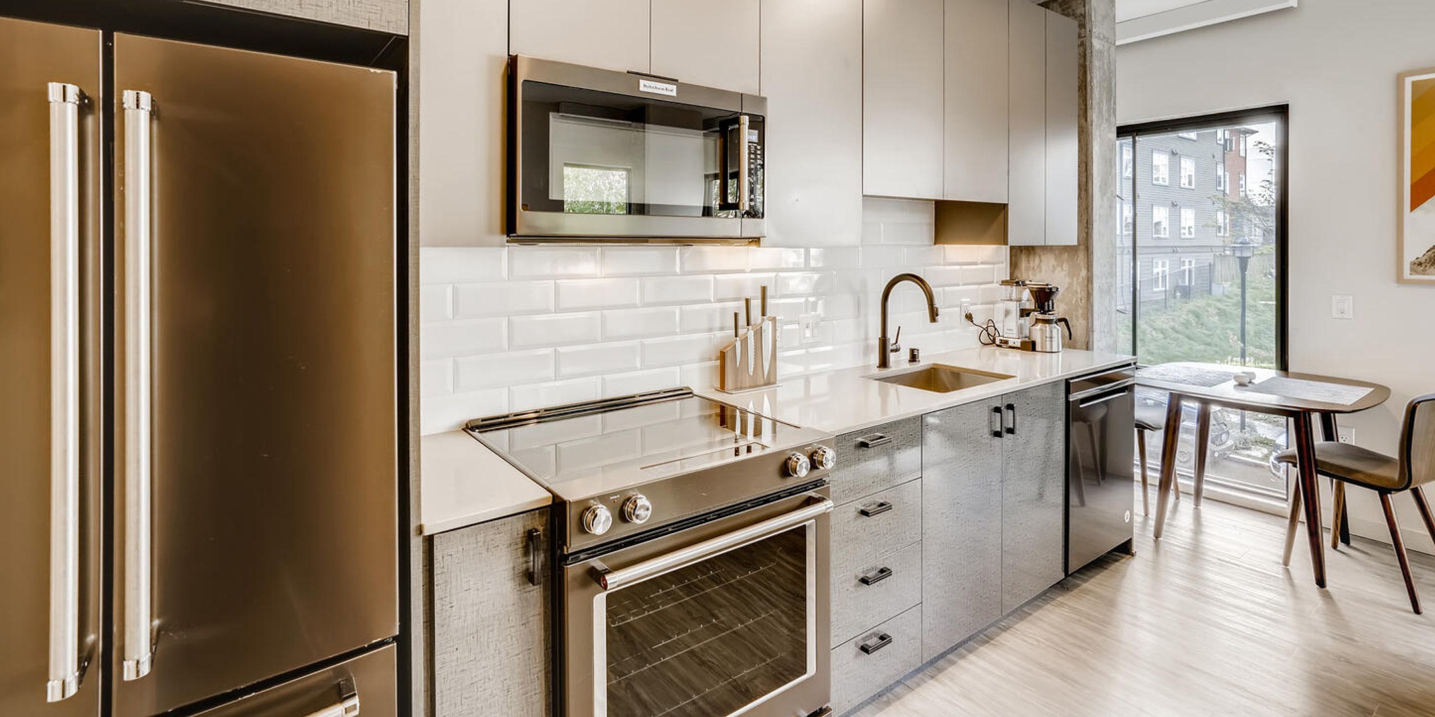 Sleek modern kitchen with bronze appliances, flat-front cabinetry, white subway tile backsplash, and a small dining table positioned beside a large window.