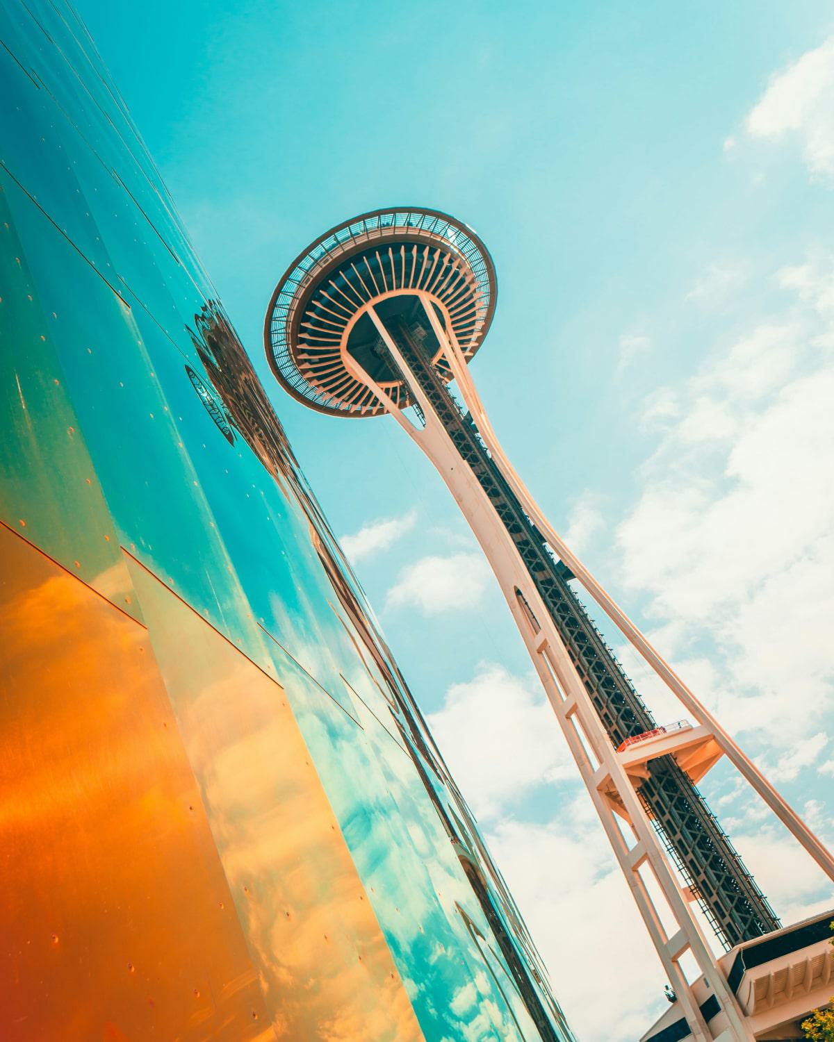 Dramatic upward view of the Space Needle in Seattle against a bright sky, with the iridescent, copper-toned exterior of the Museum of Pop Culture reflecting the clouds and sky.