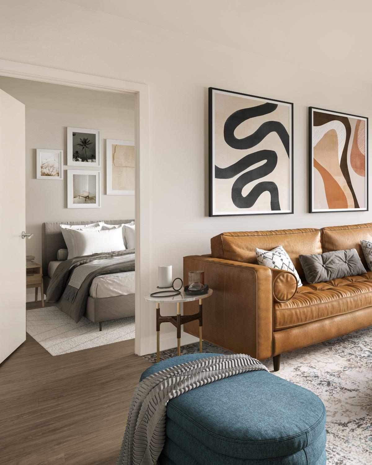 Stylish apartment living room with a caramel leather sofa, abstract wall art, and a blue ottoman, leading into a bedroom with layered bedding and framed artwork above the bed.