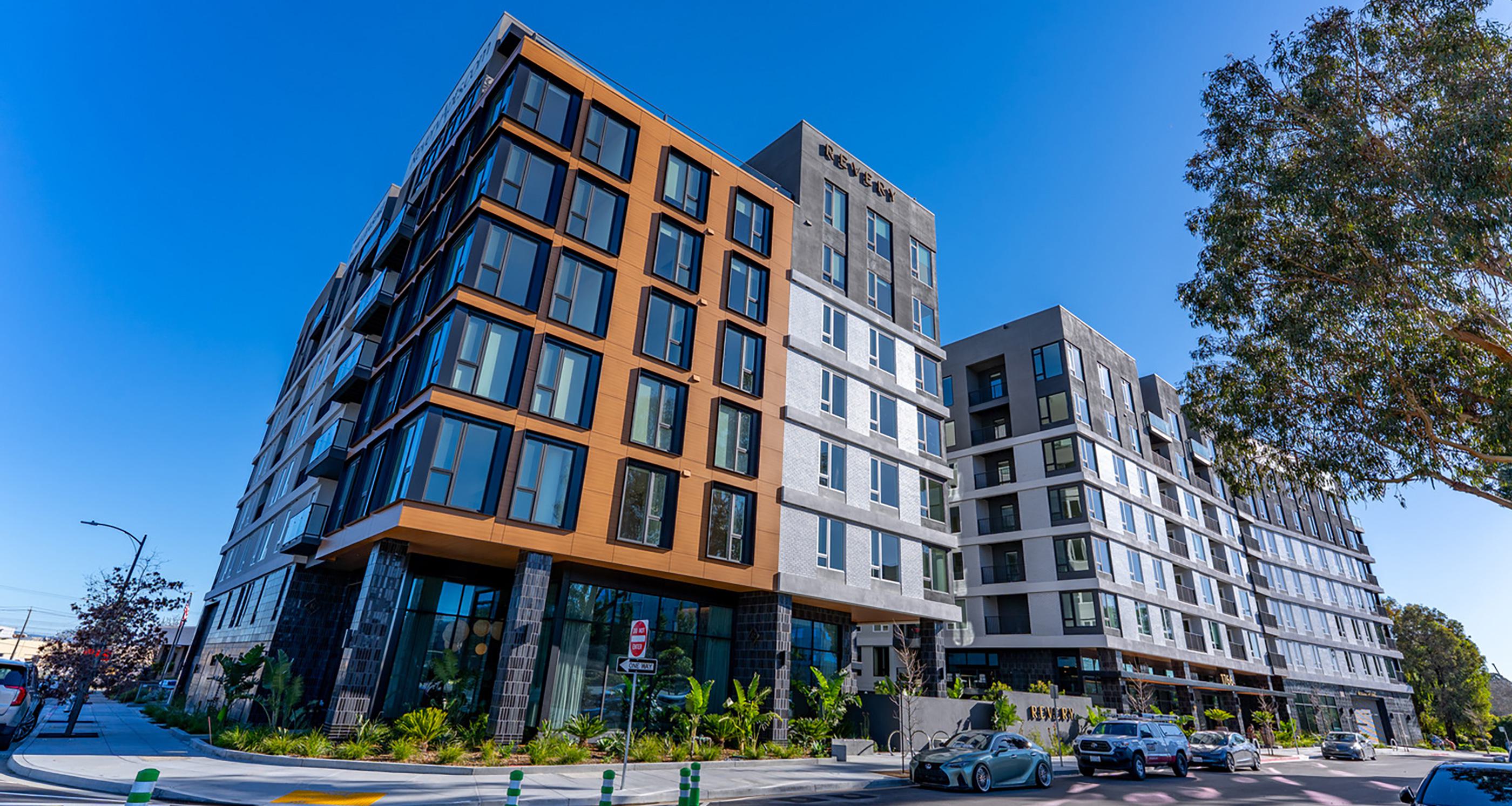 Modern,  mid-rise Revery apartment building at dusk, with sleek glass corner windows, warm interior lighting, and the name “REVERY” illuminated at the top. 