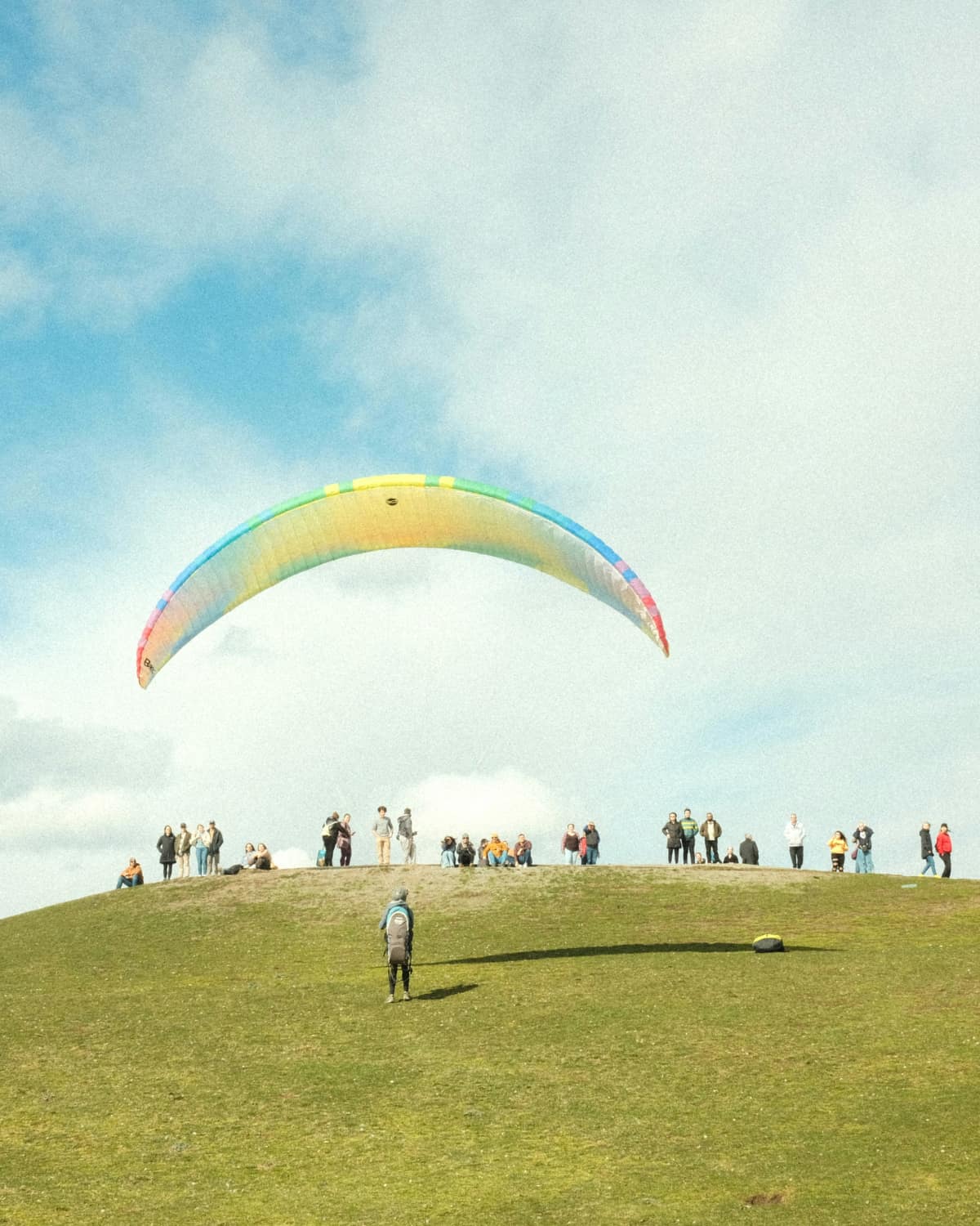 A group of people standing on top of a lush green hillside. A paraglider stands on the hillside in front of them, their rainbow parachute flying in the sky above.