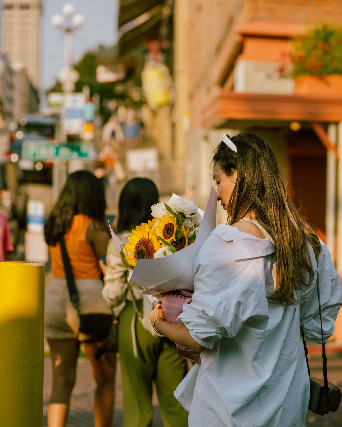A woman faces away from the camera, holding a bouquet of sunflowers on a sunny city street.