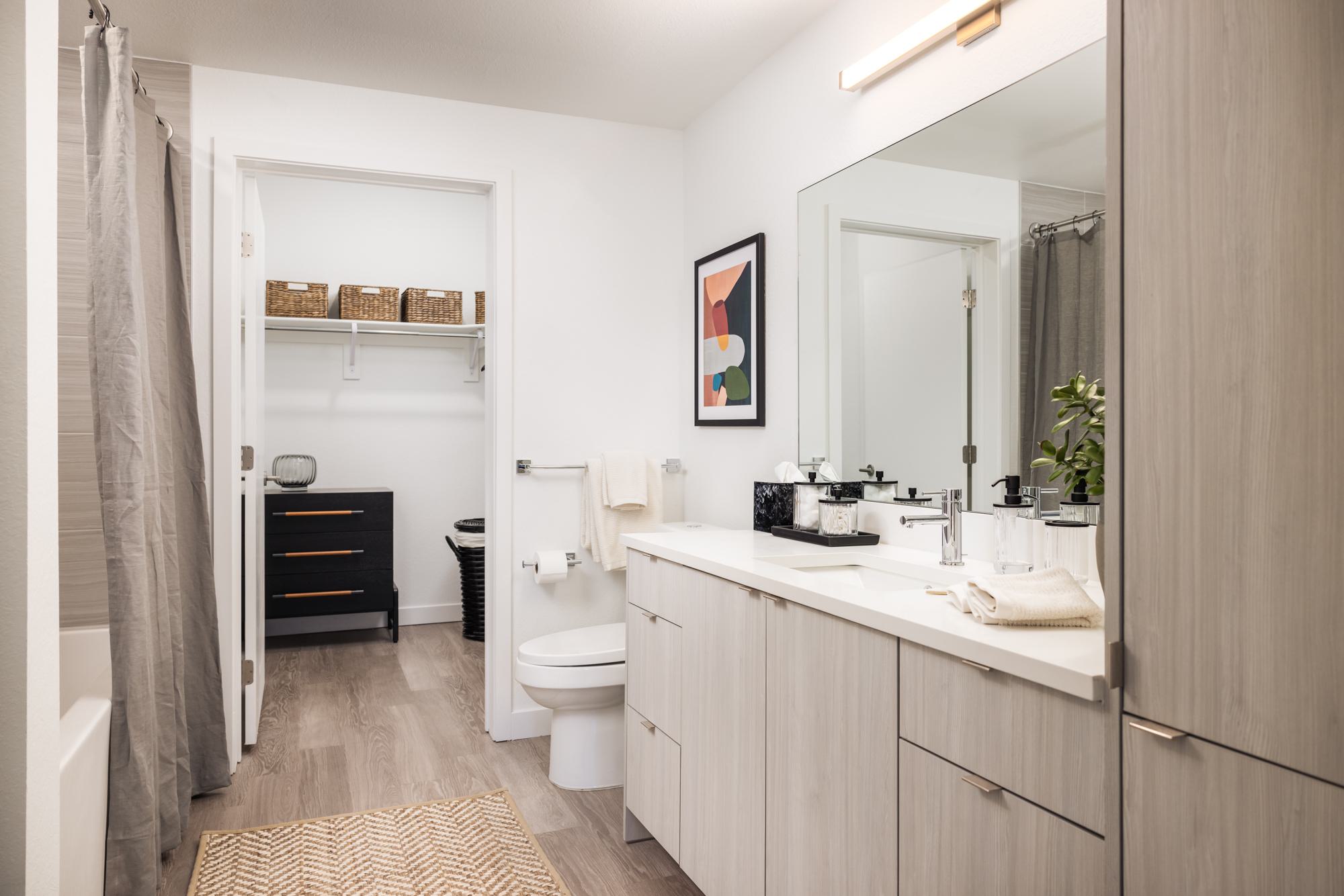 Modern bathroom at Forge Apartments in RiNo, Denver, featuring light wood cabinetry, a large mirror, soft neutral tones, and direct access to a spacious walk-in closet with built-in shelving and storage.