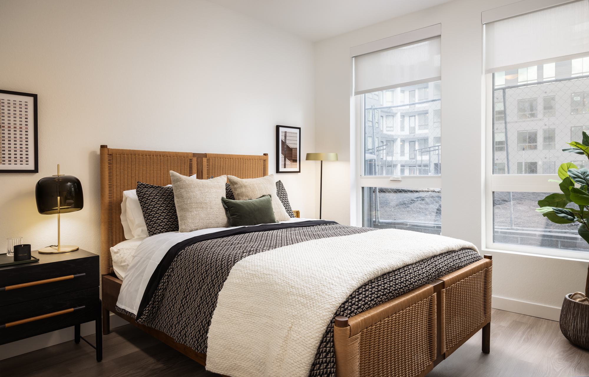 Stylish bedroom at Forge Apartments in RiNo, Denver, featuring a woven bed frame, layered neutral bedding, modern nightstand, and large windows letting in natural light.