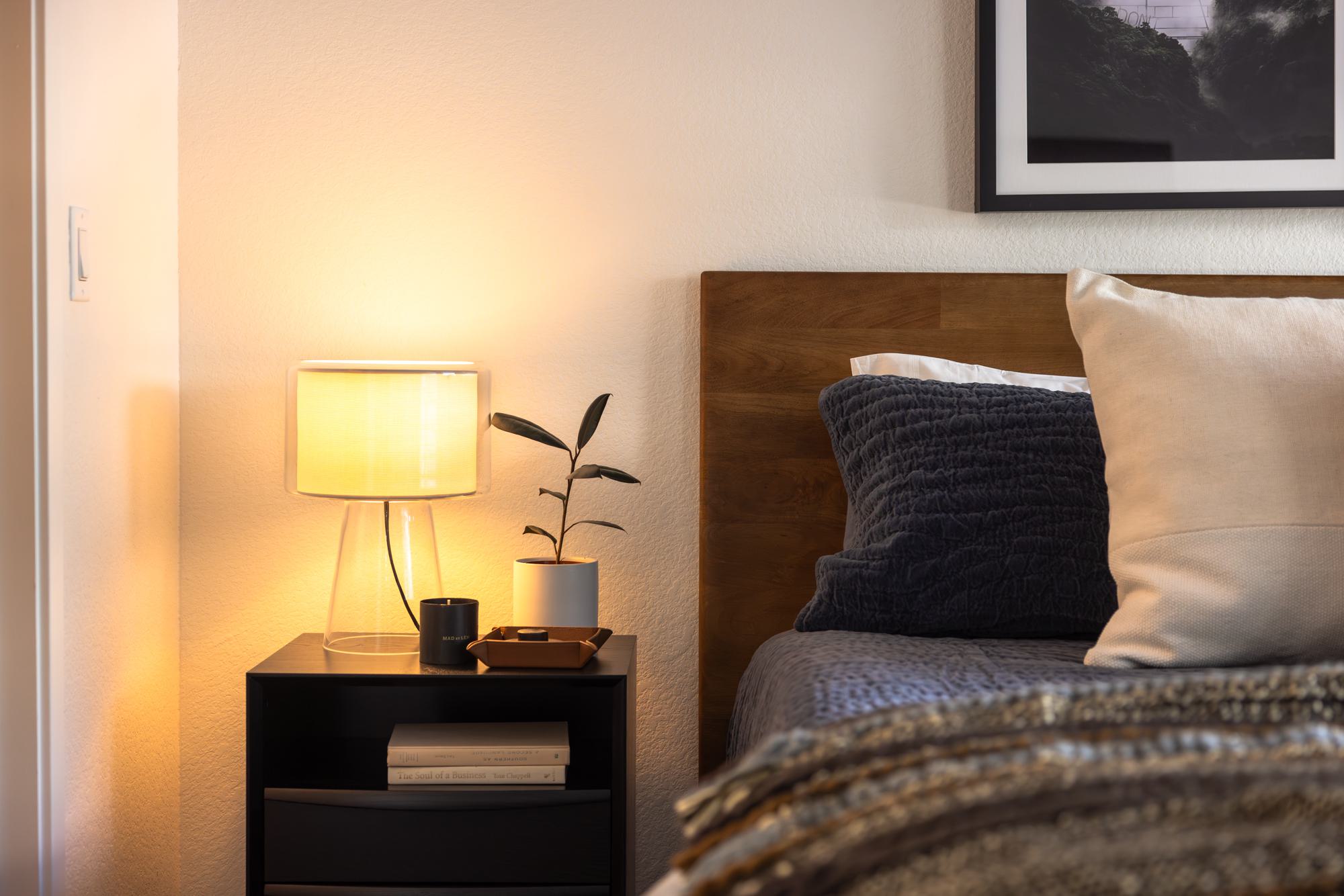 Close-up of a modern bedroom nightstand with a glowing glass lamp, potted plant, books, and a wooden headboard partially in view.