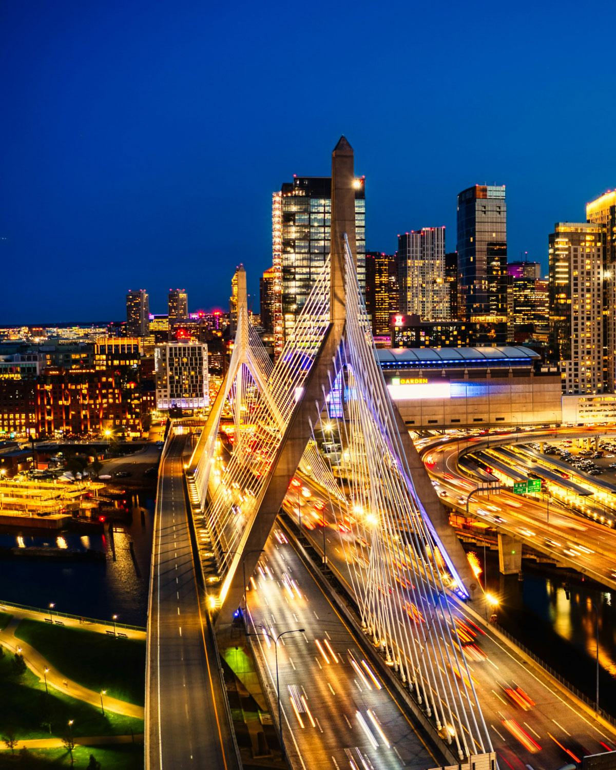 Night view of the Leonard P. Zakim Bunker Hill Memorial Bridge in Boston, illuminated with colorful lights and surrounded by city skyscrapers and traffic trails.