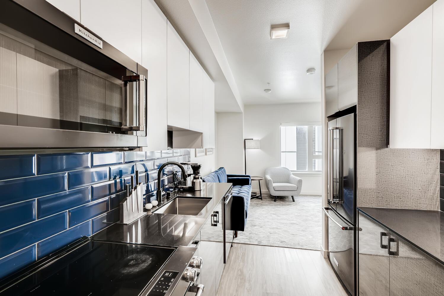 Contemporary galley kitchen with glossy black countertops, a bold navy blue tile backsplash, white upper cabinets, and bronze appliances, opening into a bright living area with modern seating.