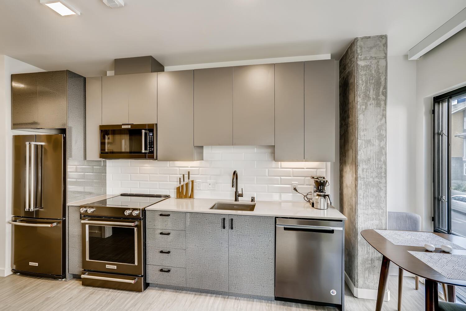  Contemporary kitchen with sleek taupe cabinetry, dark bronze appliances, white subway tile backsplash, and a compact dining table beside a large window.