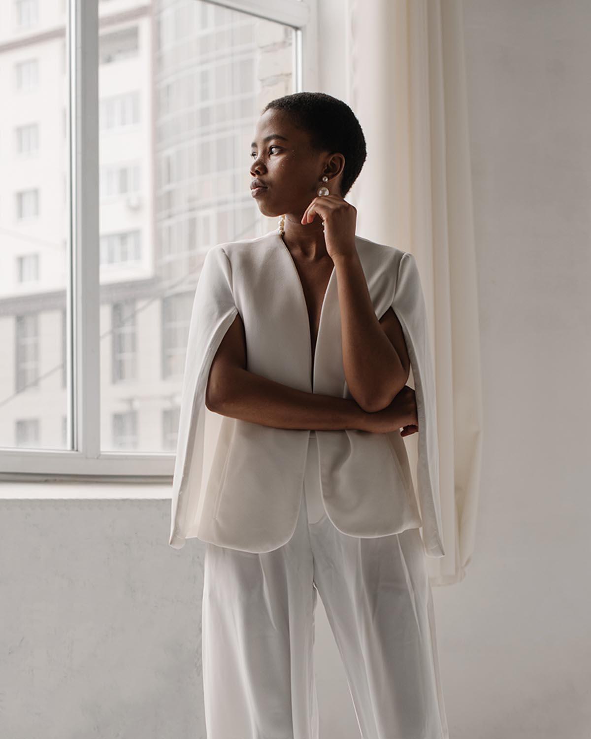 A poised woman in an elegant white cape blazer and trousers gazes thoughtfully out of a large window, framed by soft natural light against a modern urban backdrop.