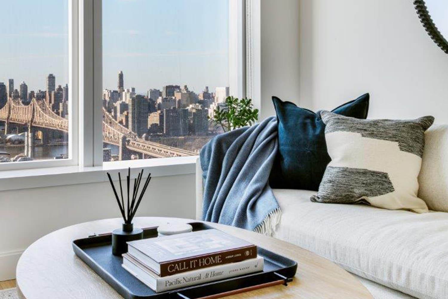 A stylish living room lounge setting features a modern wood coffee table adorned with books about home design, an aroma diffuser and sleek black tray, overlooking the Brooklyn Bridge in a an intimate and elegant ambiance with bright lighting and warm furniture