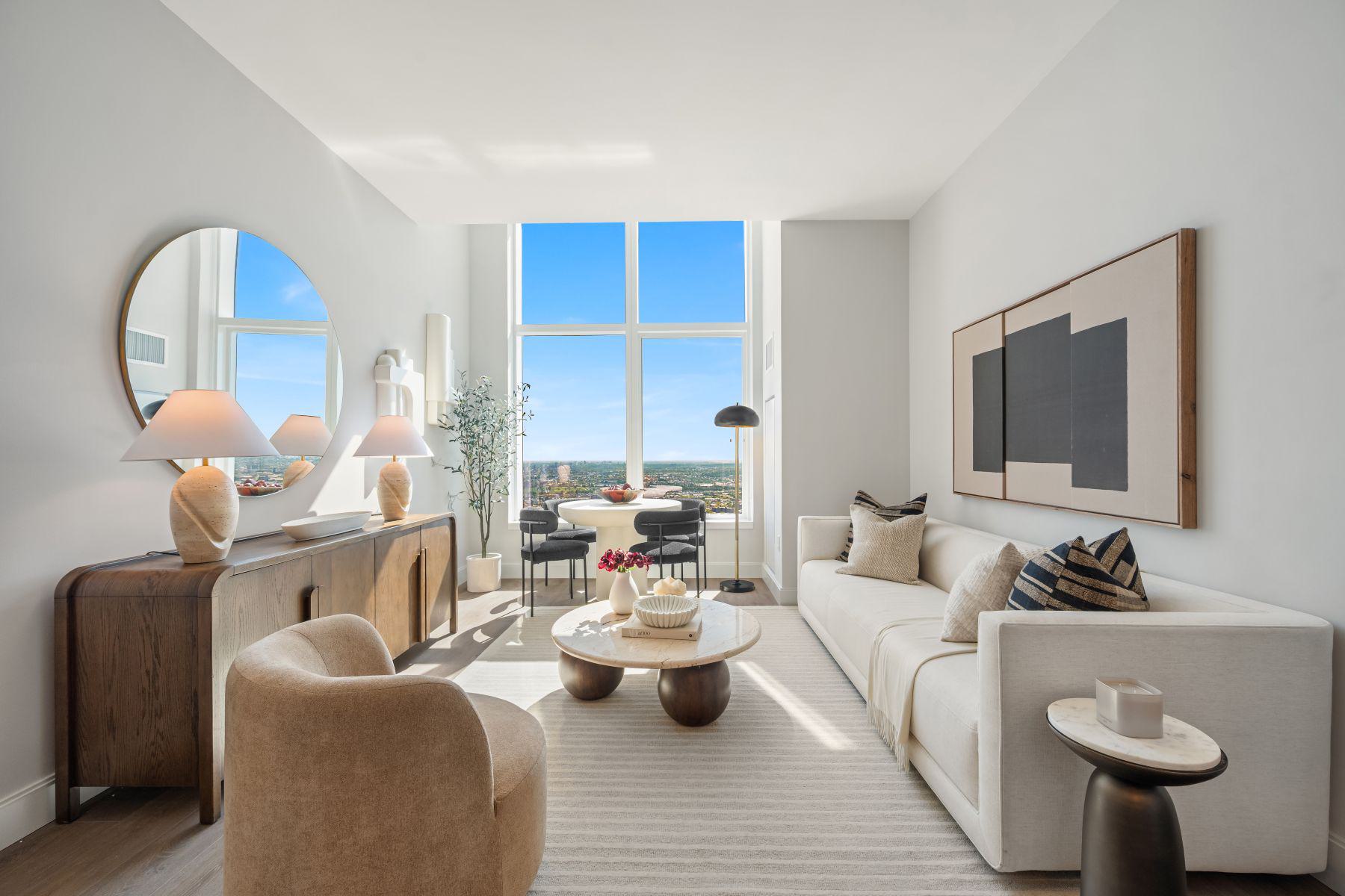 A stylish living room lounge features a sculptural marble coffee table adorned with design books, a decorative bowl and fresh flowers, complemented by warm wood accents, neutral furnishings and elegant lighting, with expansive city views creating a bright, intimate and sophisticated ambiance.