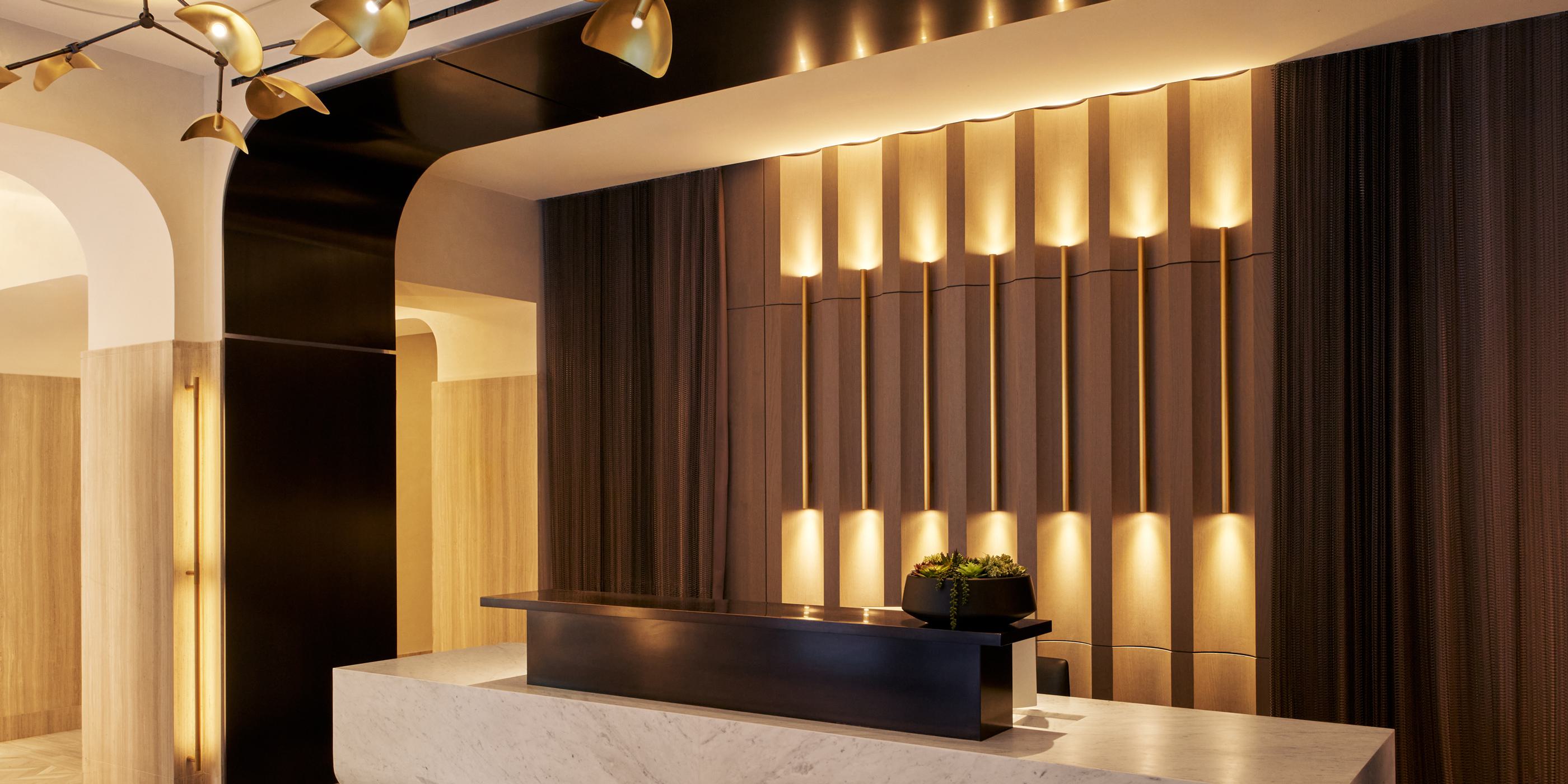 Elegant concierge lobby with a curved marble reception desk, warm wood and brass accents, and a sculptural chandelier resembling clustered glass petals. A concierge stands behind the desk in a sophisticated, modern interior.