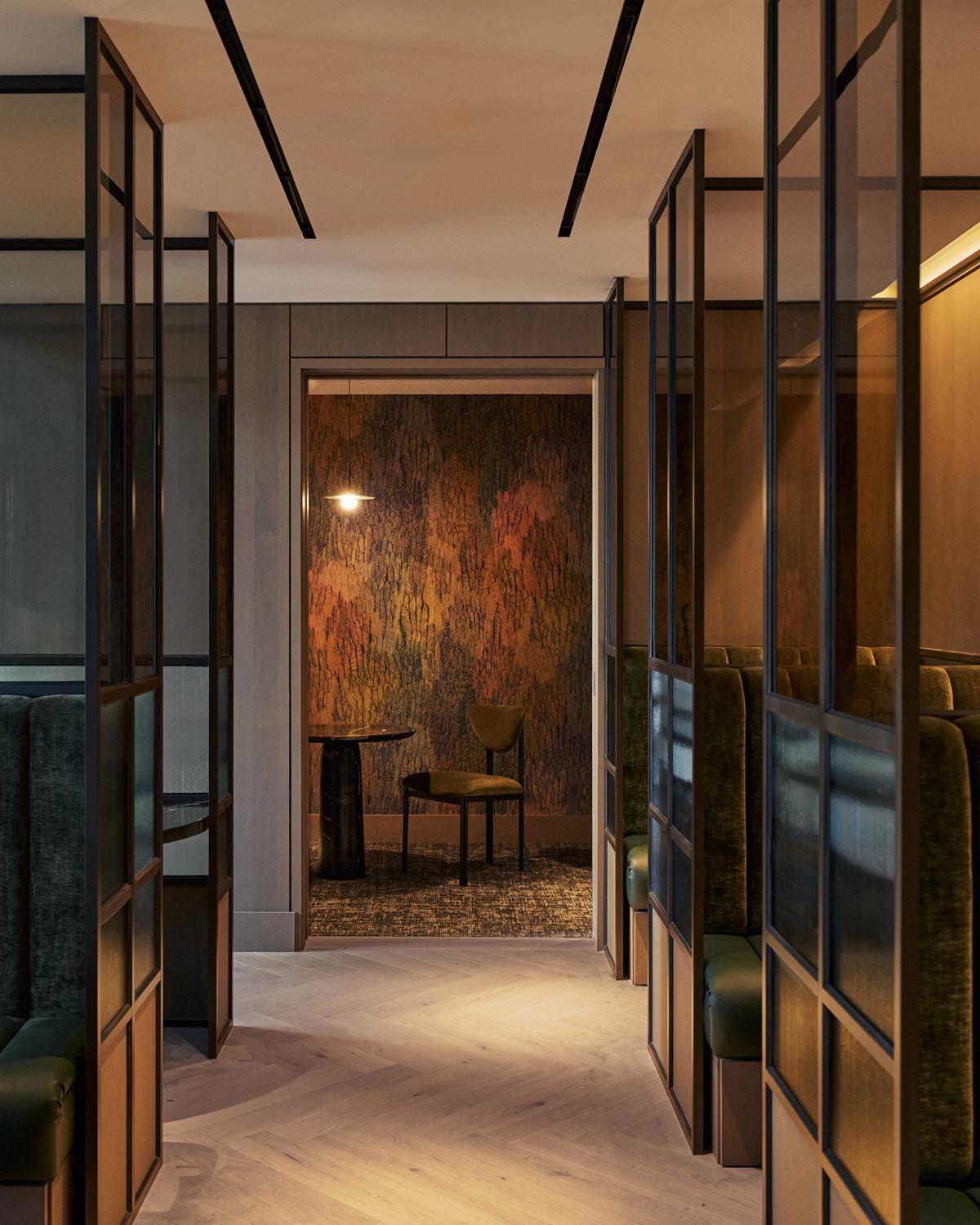 Stylish coworking lounge at Maison 78 with curved cream sofas, a marble coffee table, abstract artwork, parquet wood floors, built-in bookshelves, and green-accented dining-style desks flanking the seating area. A glass-enclosed meeting room sits at the center of the space.