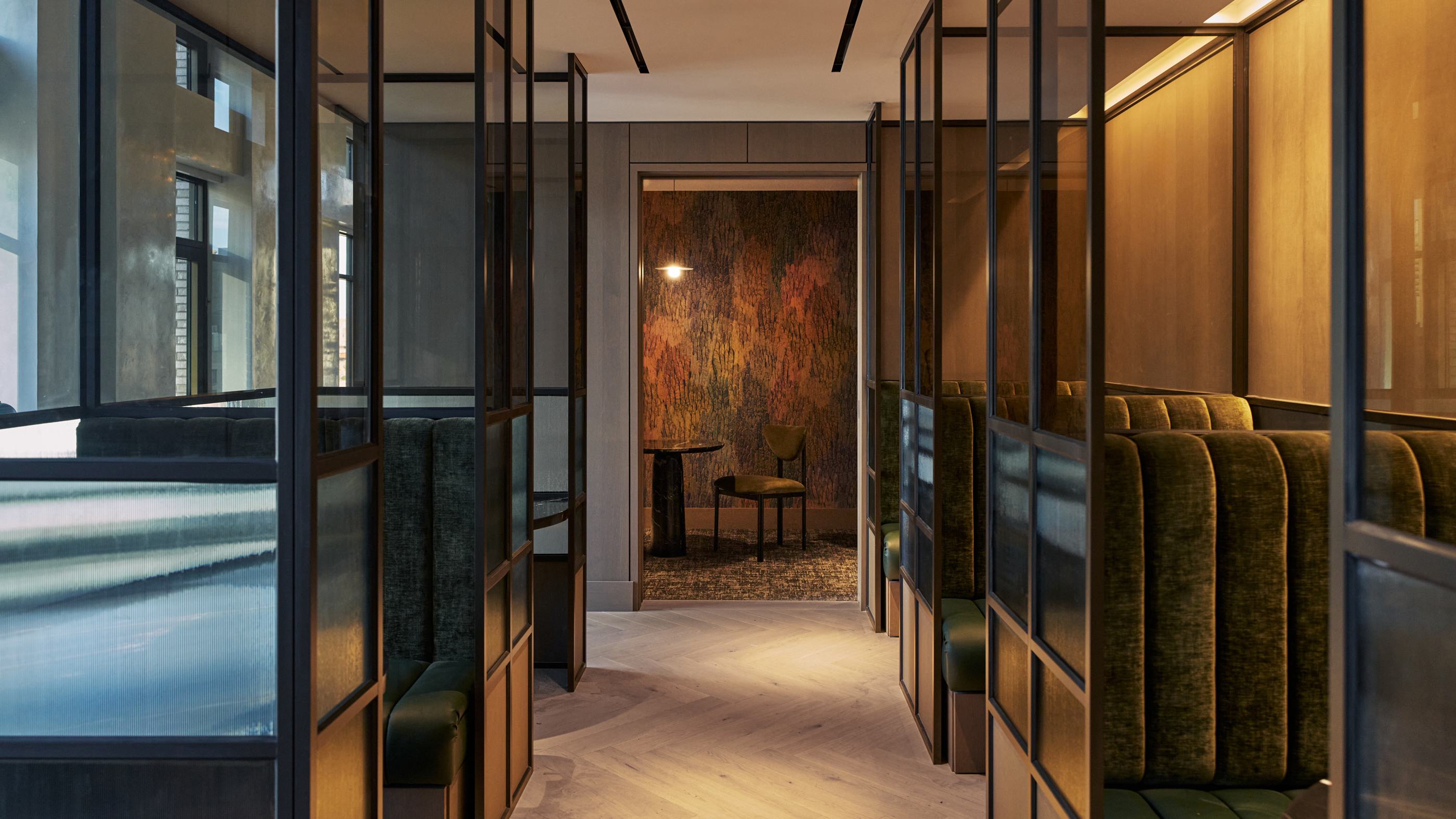 Stylish coworking lounge at Maison 78 with curved cream sofas, a marble coffee table, abstract artwork, parquet wood floors, built-in bookshelves, and green-accented dining-style desks flanking the seating area. A glass-enclosed meeting room sits at the center of the space.