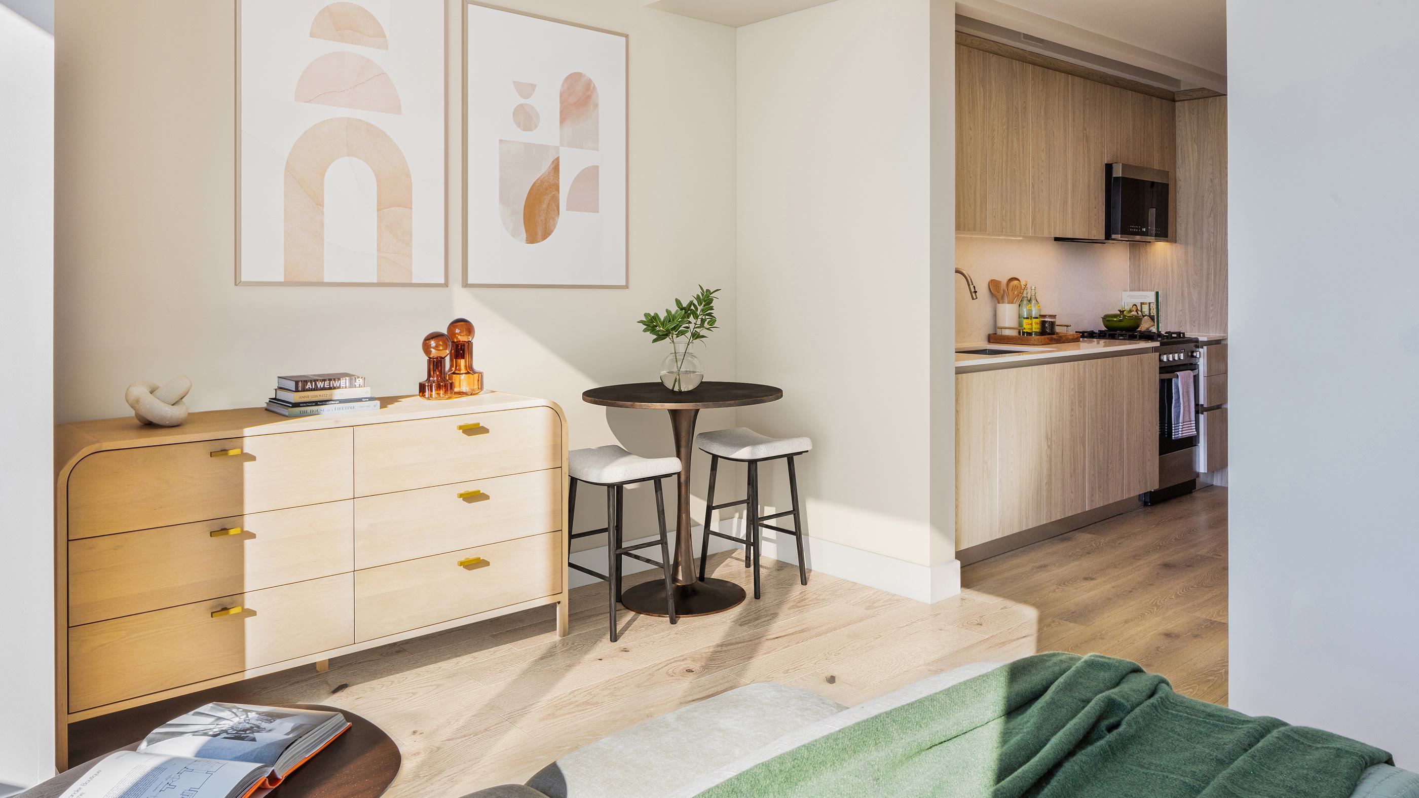 A modern open-concept kitchen and dining area with floor-to-ceiling windows, warm wood accents, sleek furnishings, and a stunning leafy views of prospect park and Manhattan skyline in the distance