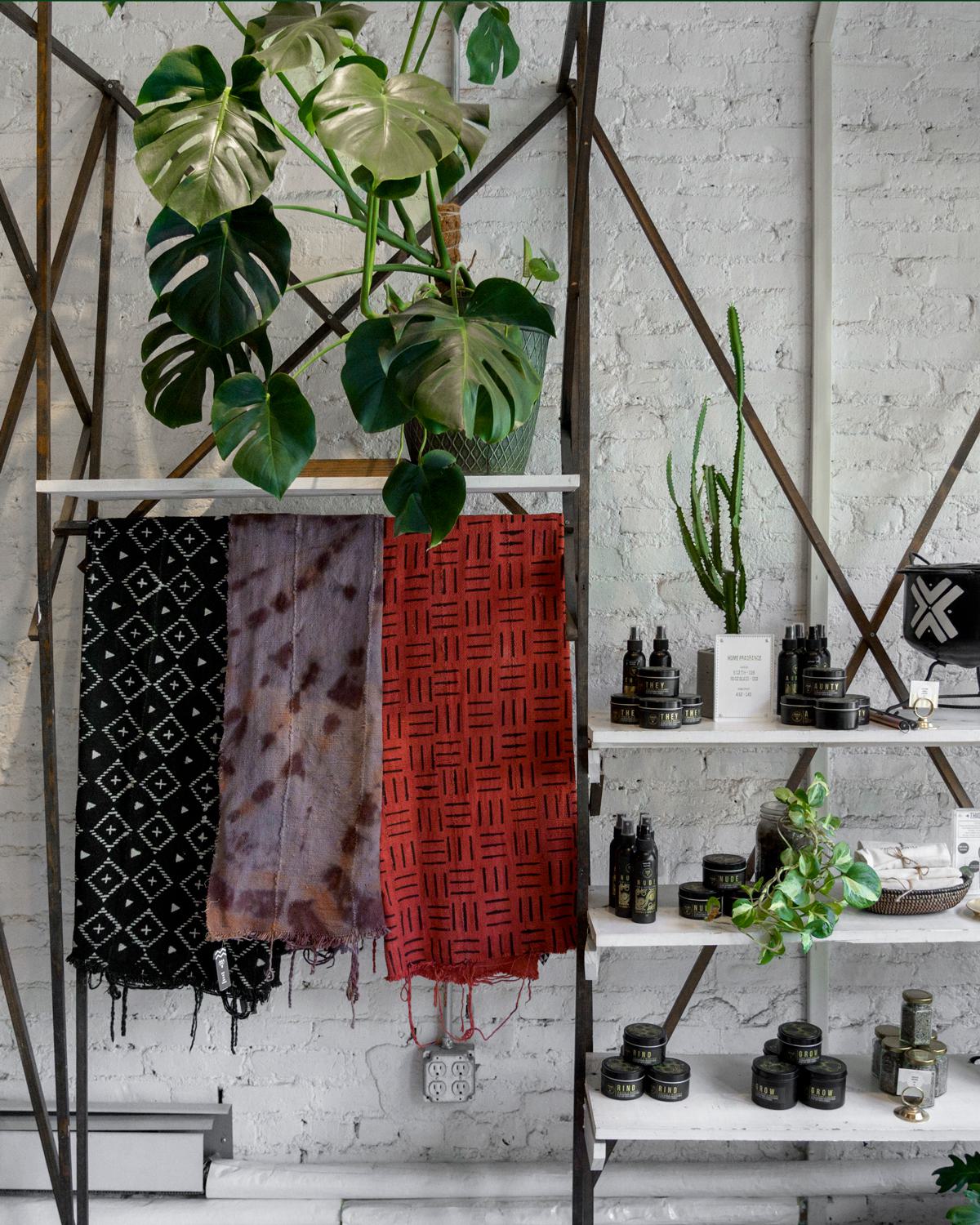 tylish boutique display featuring handcrafted textiles, natural skincare products, and lush monstera plans and greenery against a white brick wall,