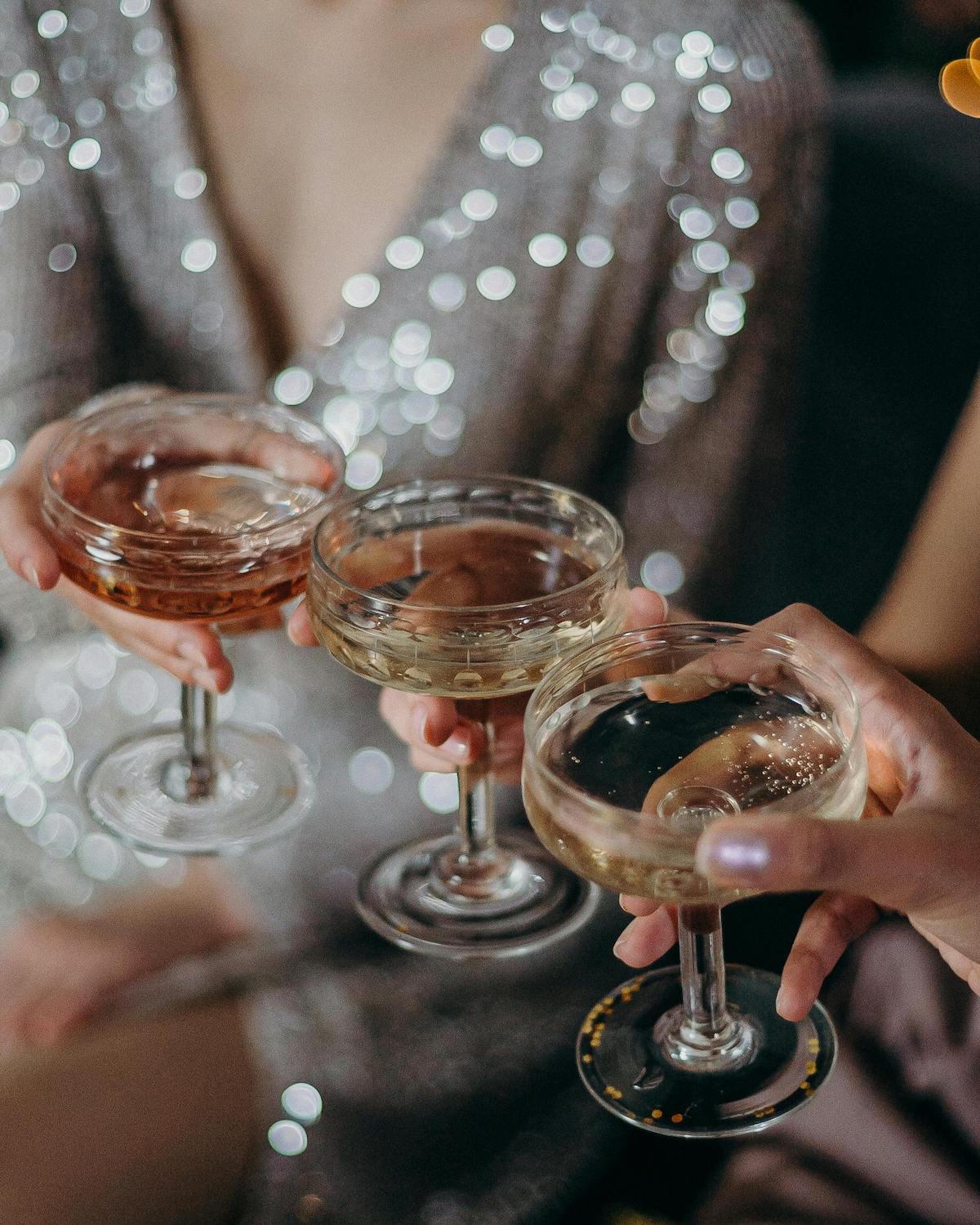 Close-up of three people clinking coupe glasses filled with sparkling wine and champagne, with one person wearing a shimmering silver sequin outfit in the background.