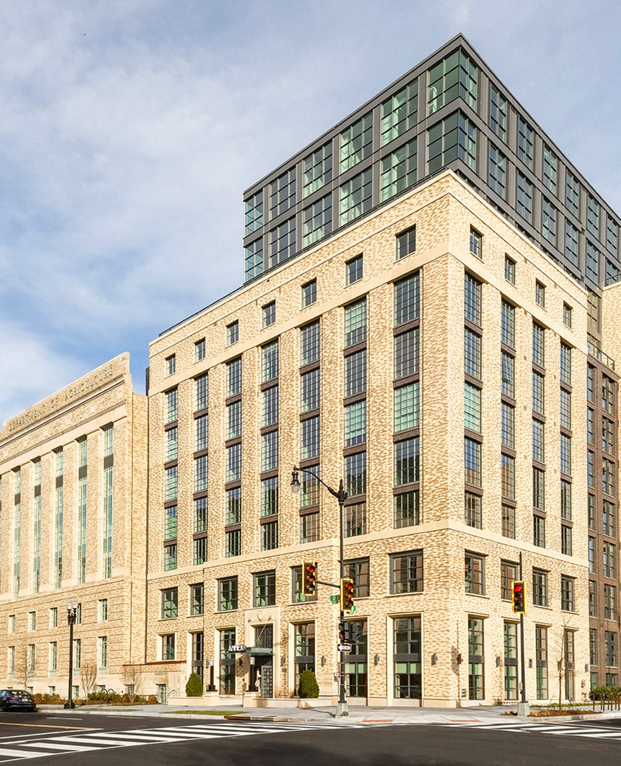 Daytime view of Annex on 12th in Southwest DC, featuring restored cream-colored brickwork, tall industrial-style windows, and a modern glass-clad upper addition above the historic Department of Agriculture façade.