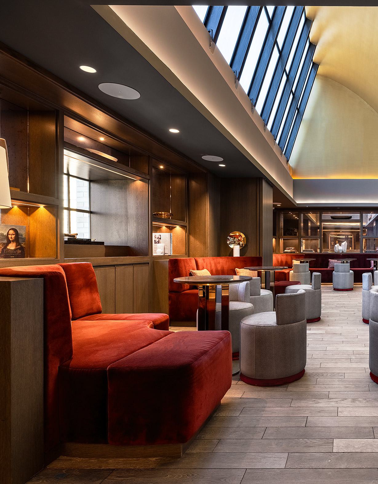 Elegant speakeasy-style lounge with plush red and gray seating, wood-paneled walls, curated bookshelf niches, and a dramatic barrel-vaulted ceiling with skylights.