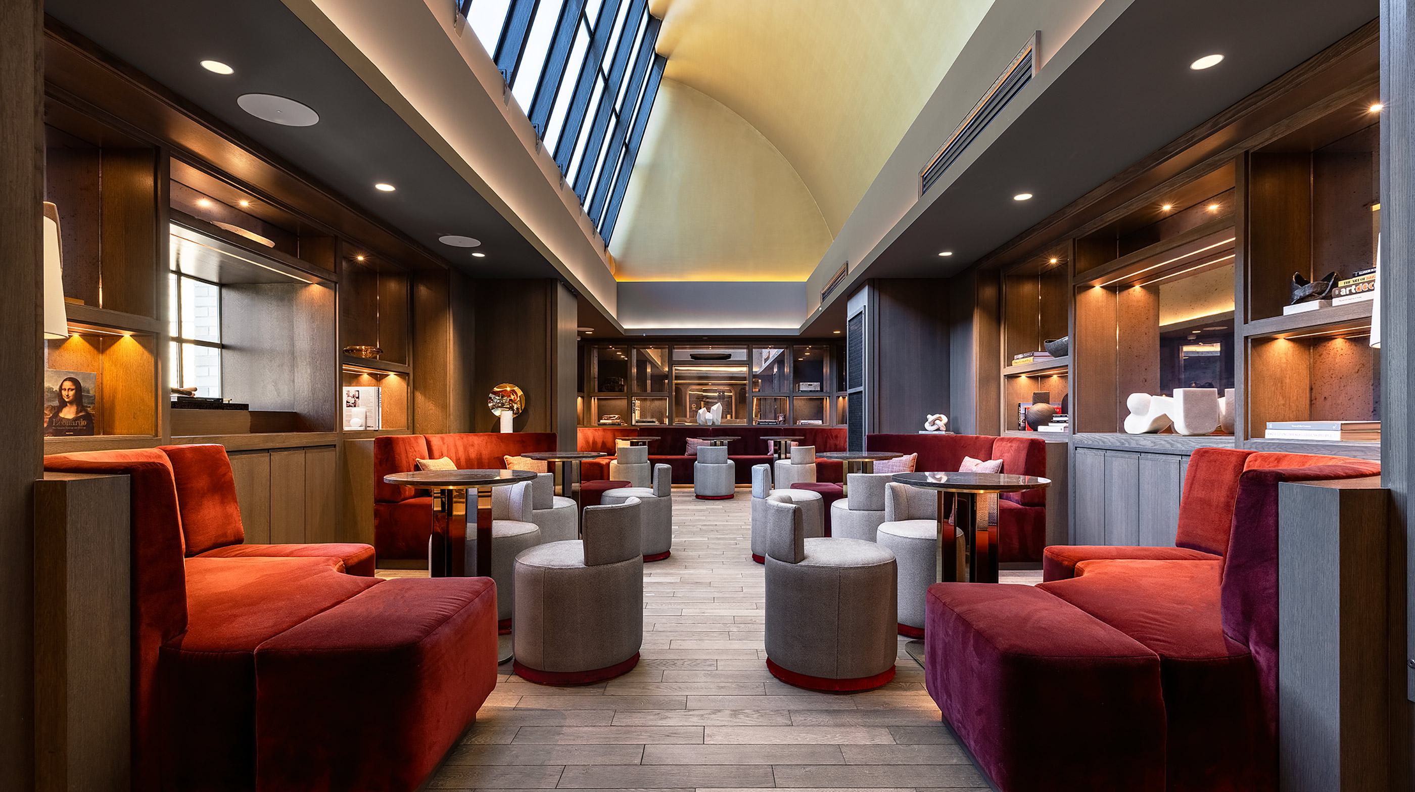 Elegant speakeasy-style lounge with plush red and gray seating, wood-paneled walls, curated bookshelf niches, and a dramatic barrel-vaulted ceiling with skylights.